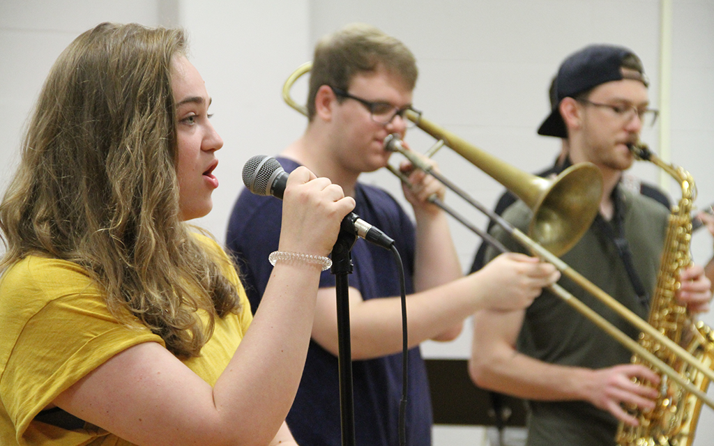 A young student in the Jefferson College Music Department sings into a microphone, with two fellow students playing horns behind her.